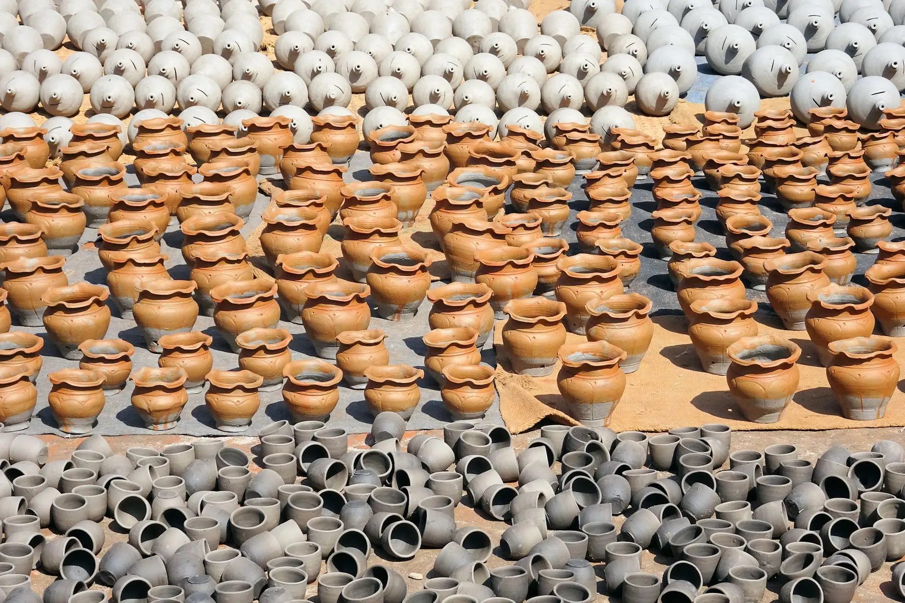 Rows of freshly made clay pots drying in the sun, orange pots in the middle and unfired grey pots in front and behind.