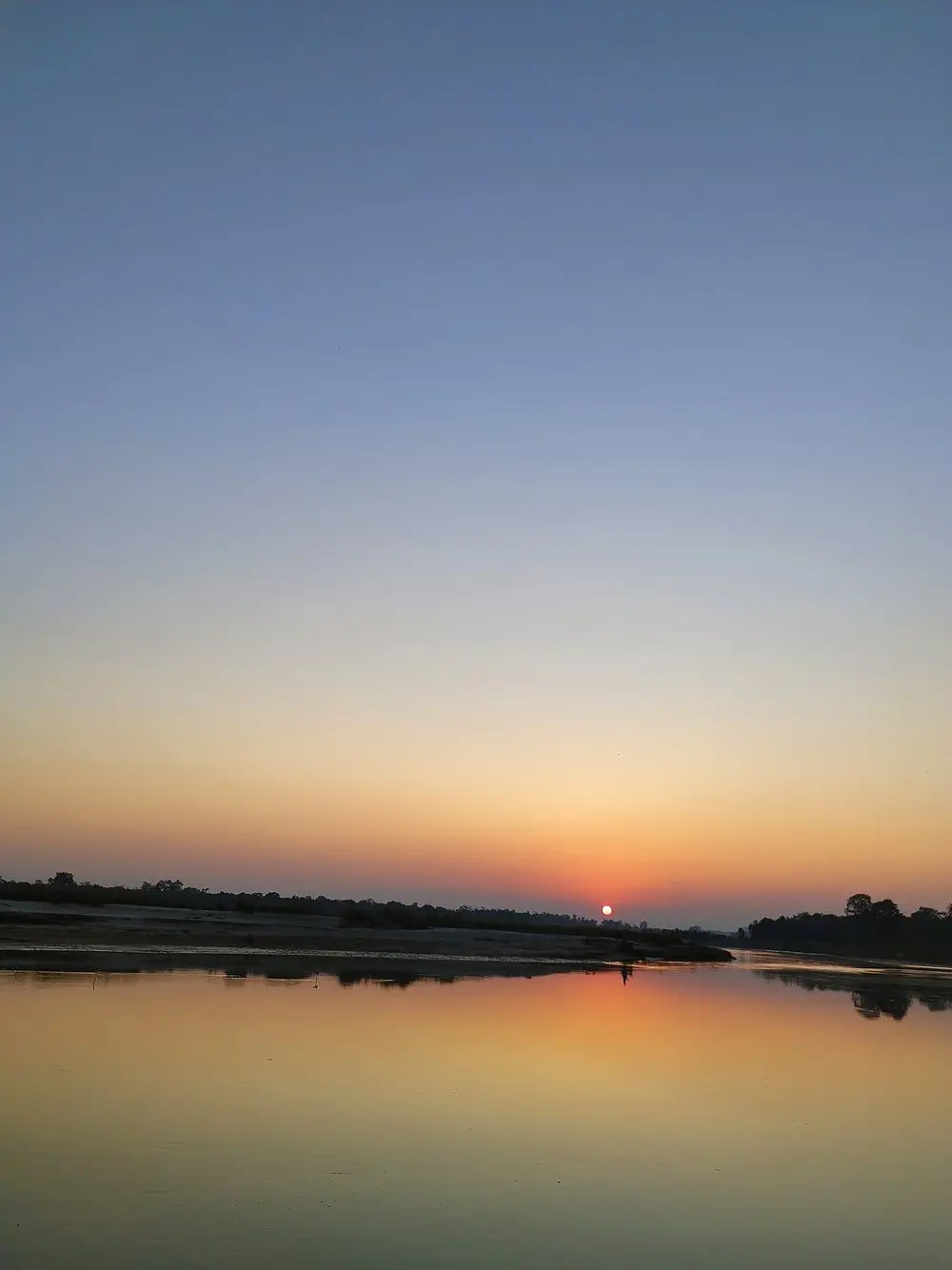 The sun setting low over the Mohana River, its glow reflected in still water.