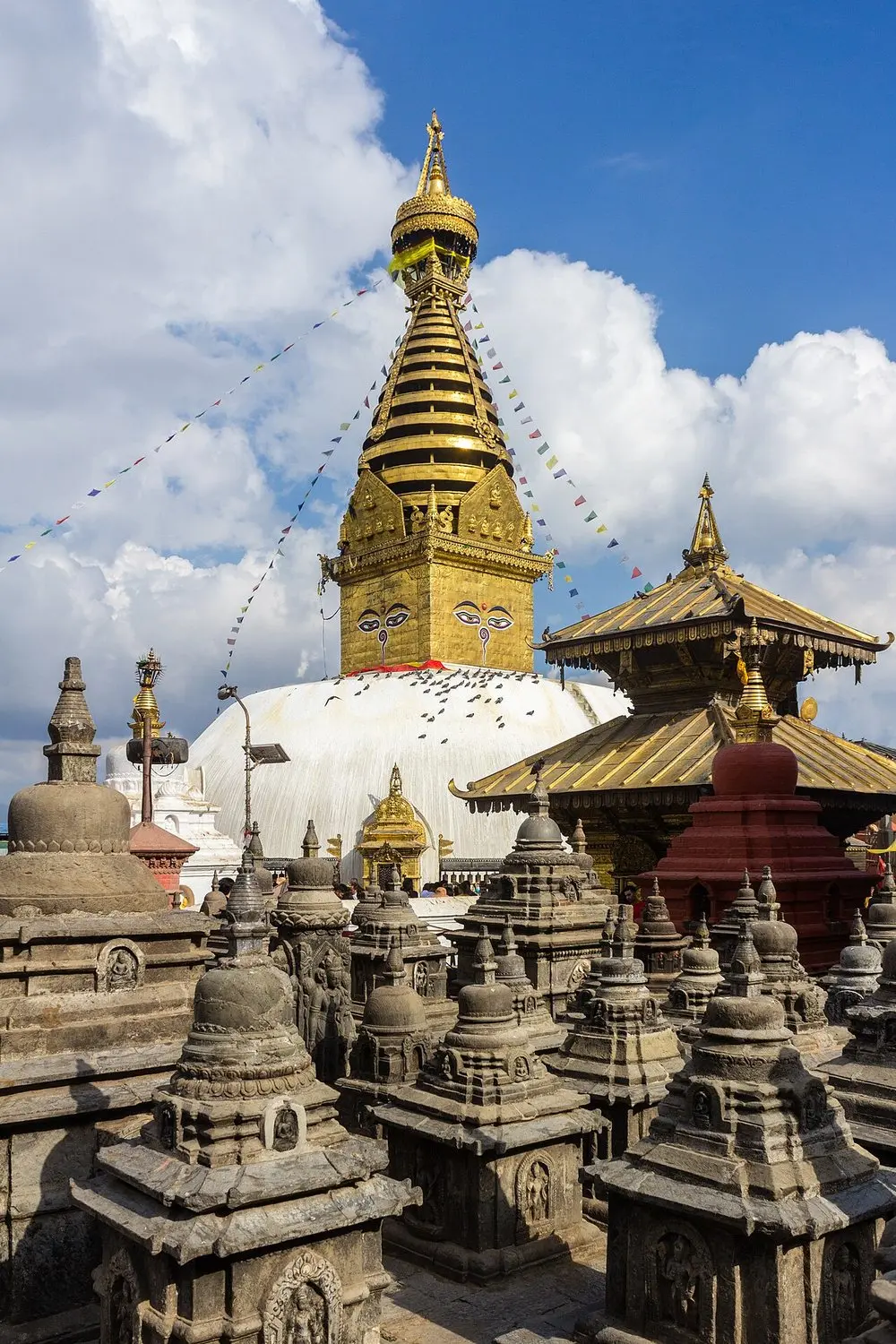 The gilded spire of Swayambhunath stupa above a cluster of small stone shrines, against a blue sky.