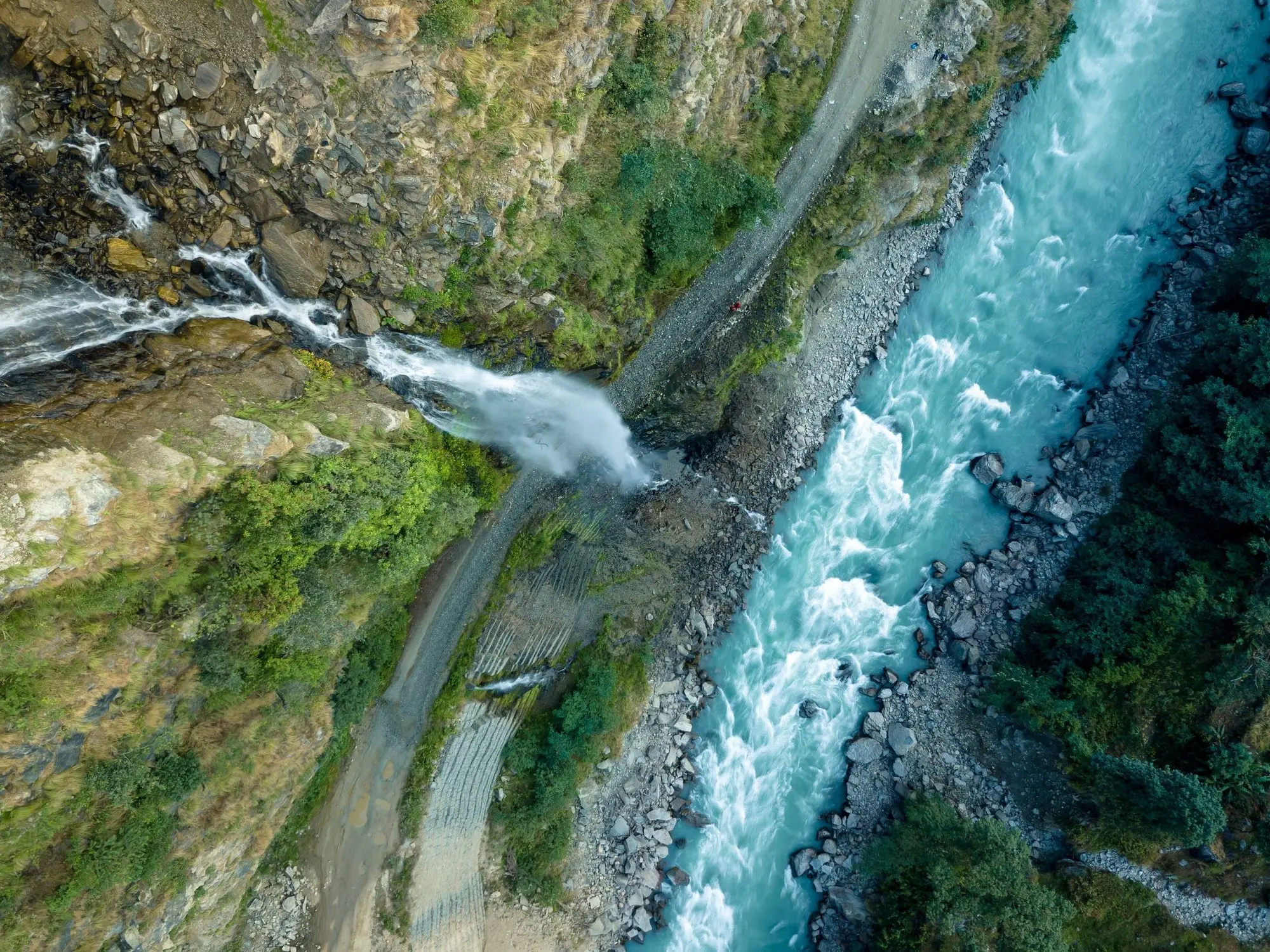 Looking straight down on a turquoise river, a waterfall joining it and a road cut along the forested bank.